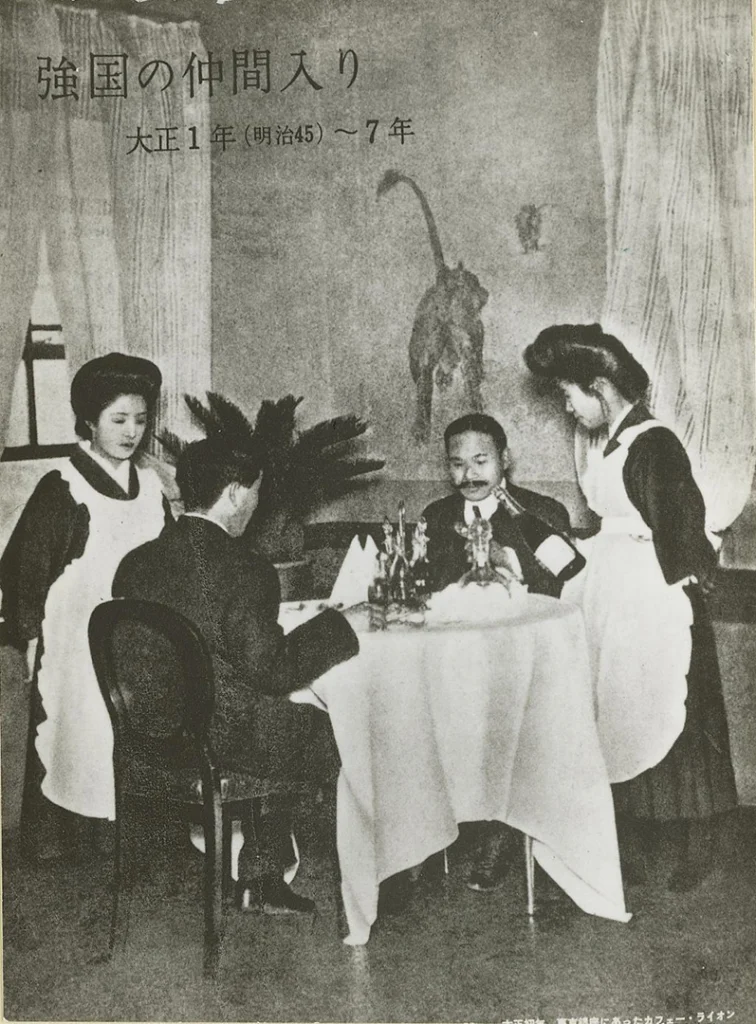 Fotografia storica in bianco e nero dell'interno del Café Lion a Ginza, Tokyo, durante l'era Taishō. Due uomini siedono a un tavolo con tovaglia bianca mentre due jokyū, che indossano il tipico mix di kimono e grembiule bianco, assistono al servizio. Sullo sfondo è visibile un murale raffigurante un leone. Collezione della Biblioteca Kyobashi.
