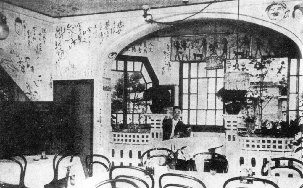 Un'immagine fotografica storica in bianco e nero dell'interno del Café Printemps a Ginza, Giappone, un pionieristico esperimento di precoce 'Kafē'. Mostra tavoli e sedie bentwood vuoti in primo piano e un arco centrale. Le pareti sono coperte da testo scritto a mano, caricature e un fregio orizzontale di figure stilizzate. Un membro dello staff sorridente è visibile dietro una balaustra vicino a porte-finestre a griglia. Questa immagine fornisce una vista dettagliata dell'ambiente unico e artistico di questo specifico caffè giapponese.
