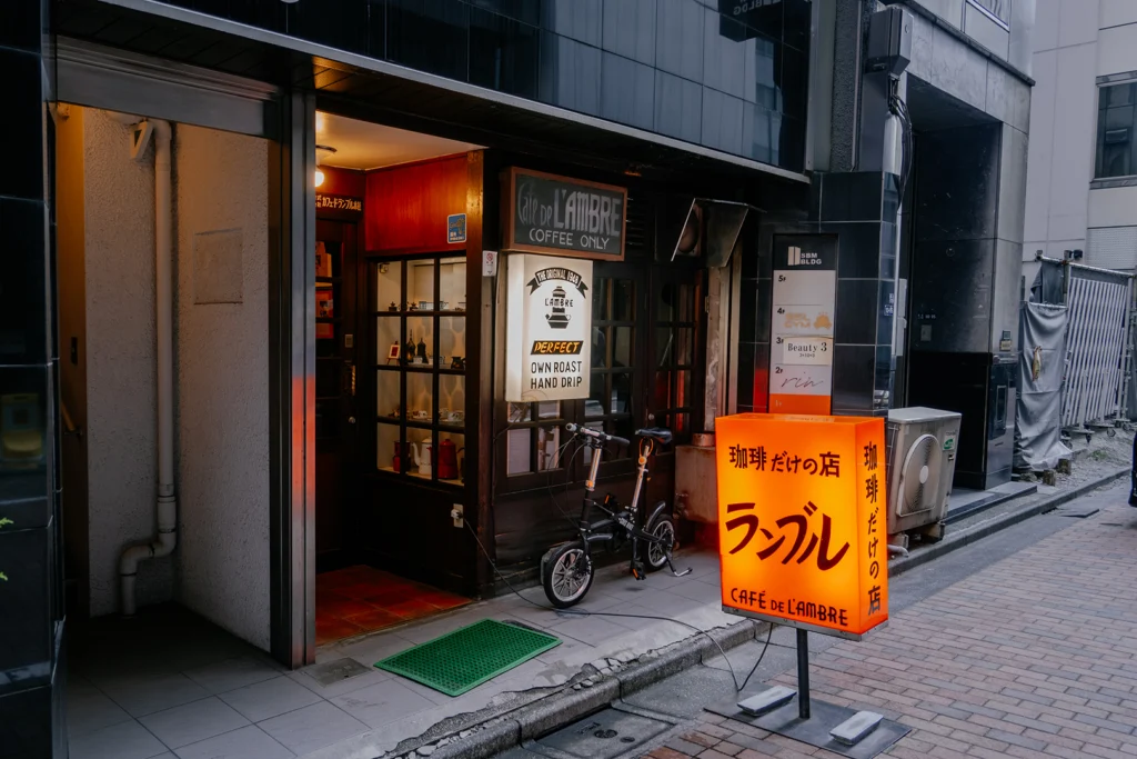 L'ingresso esterno dello storico kissaten Café De L'Ambre a Ginza, Tokyo. Si vedono le insegne vintage in legno 'Coffee Only' e l'iconica insegna luminosa arancione lungo una caratteristica via cittadina.