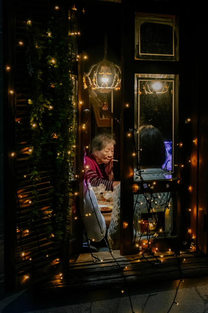 Scorcio notturno del Kissaten Kissa Kishi a Kanazawa: una finestra decorata con luci soffuse mostra un cliente all'interno, rappresentando il kissaten come un rifugio intimo e silenzioso.