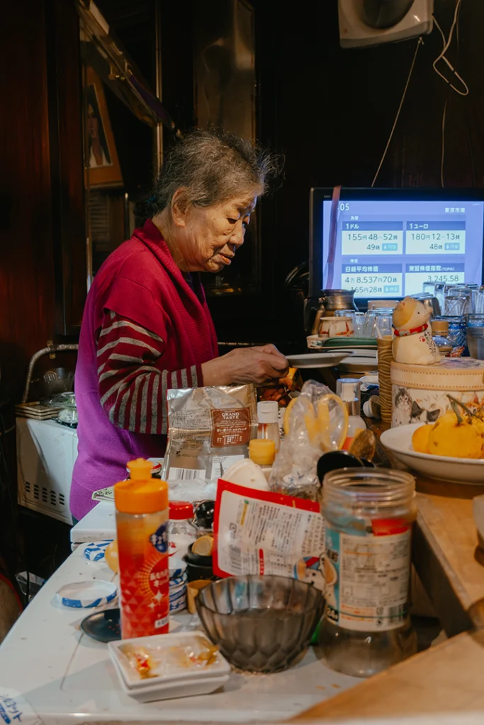 L'anziana proprietaria del Kissaten Kissa Kishi a Kanazawa mentre lavora dietro il bancone colmo di oggetti quotidiani, catturando l'autenticità e la dedizione dei caffè tradizionali giapponesi.