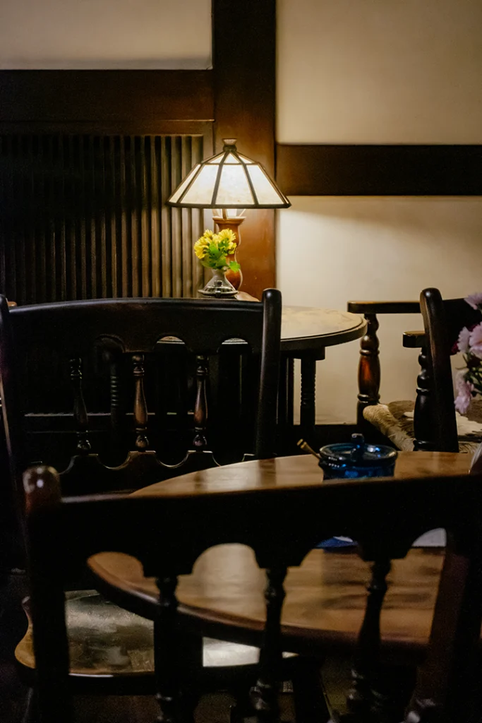 Scorcio del Ranka Retro Café a Takayama, un esempio di kissaten tradizionale con mobili in legno scuro, atmosfera silenziosa e una lampada classica che crea un'illuminazione ambrata in stile Shōwa.