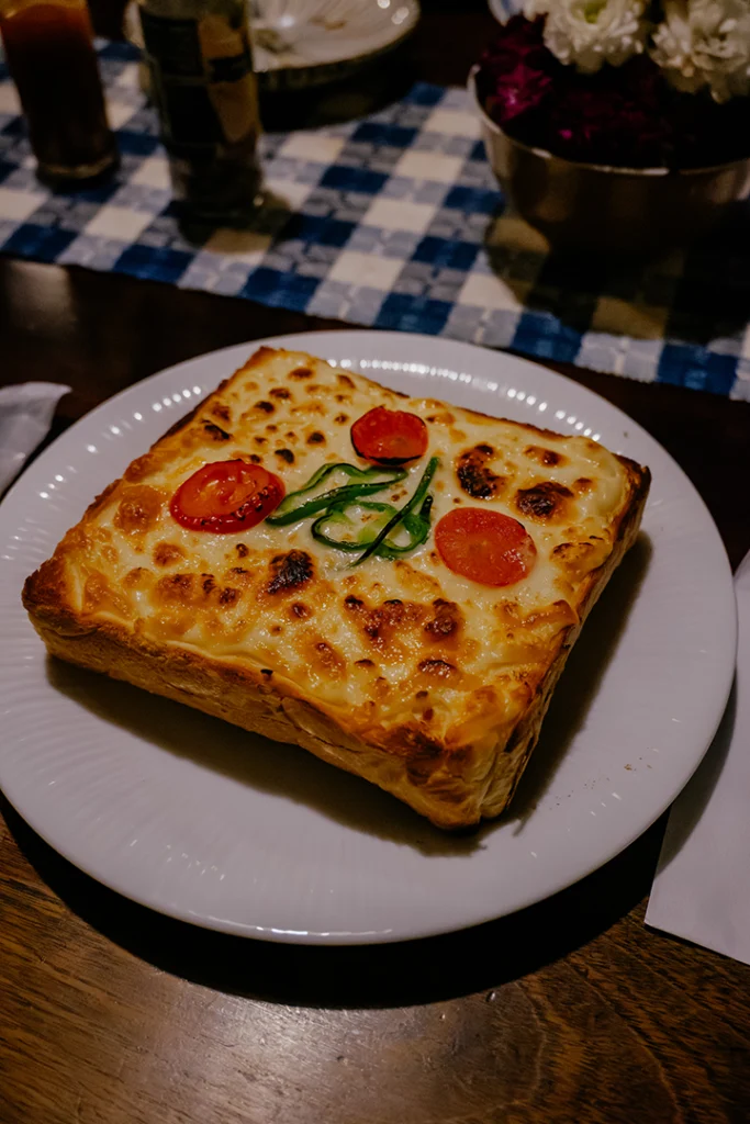 Una fetta alta di morbido pane shokupan servita come Pizza toast, un piatto classico della cucina yōshoku nei kissaten giapponesi, condita con formaggio fuso dorato, pomodorini e peperoni.