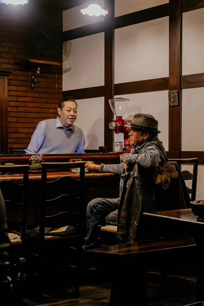 Il Master del Ranka Retro Café a Takayama conversa con un cliente al bancone in legno, mostrando l'atmosfera intima e il senso di comunità di un kissaten tradizionale giapponese.
