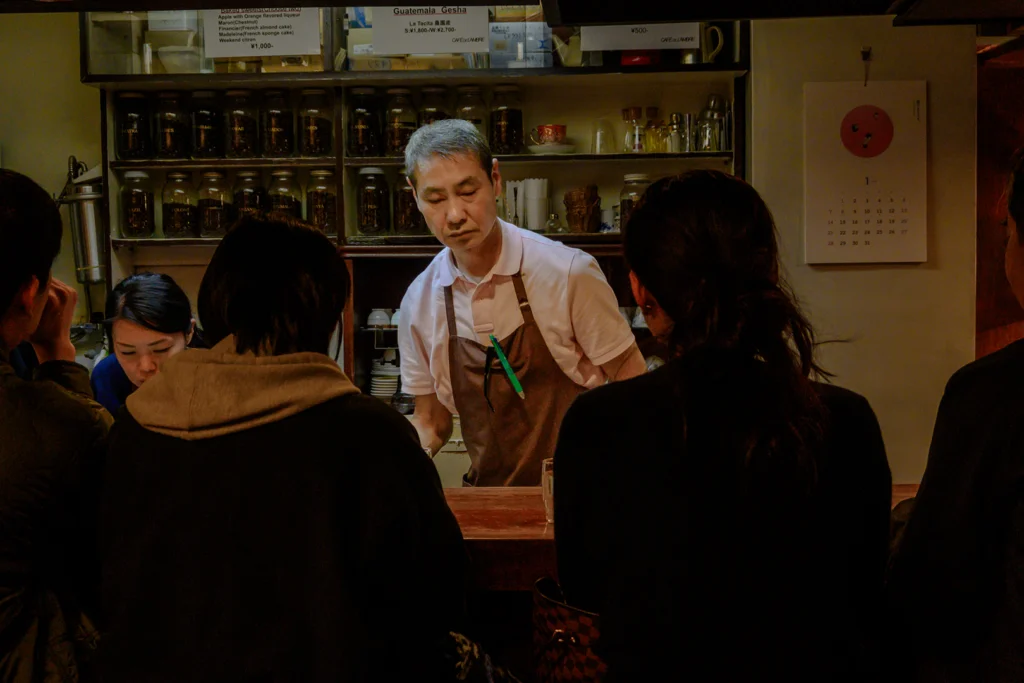 Il Master del celebre Café De L'Ambre a Ginza, Tokyo, concentrato nel servire i clienti al bancone, circondato da barattoli di chicchi di caffè in un ambiente autentico Shōwa Retro.