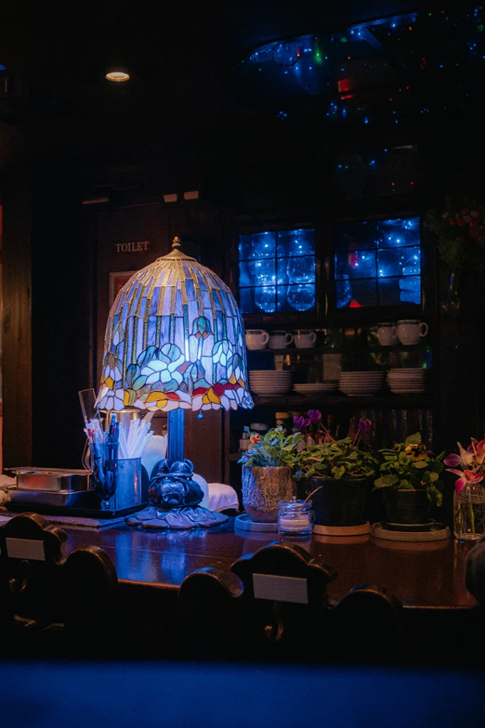 Dettaglio dell'arredamento del Café Gion ad Asagaya, Tokyo: una lampada in stile Tiffany illumina il bancone in legno scuro, creando l'atmosfera ambrata e protetta tipica di un jun kissaten.