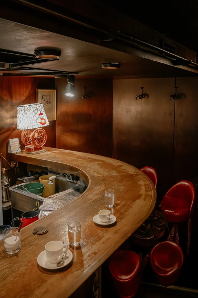 Interno dello storico kissaten Café de L'ambre a Ginza, Tokyo. Si vede l'iconico bancone in legno curvo con tazze di caffè in porcellana bianca e sedie rosse vintage, illuminato da una lampada soffusa.
