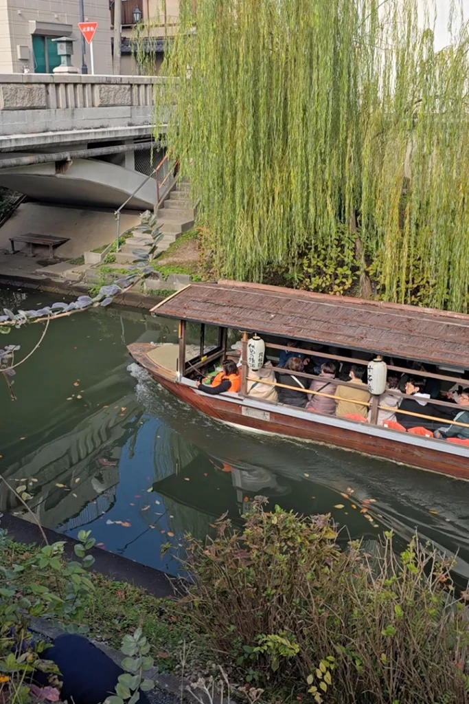 La barca tradizionale in legno Jikkokubune con turisti a bordo mentre naviga sul canale di Fushimi a Kyoto, sotto grandi salici piangenti e un ponte in pietra.