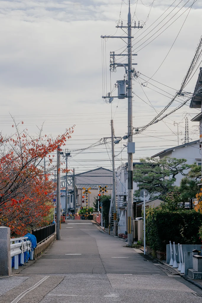Una strada secondaria a Fushimi, Kyoto, che conduce verso un passaggio a livello ferroviario con le caratteristiche luci rosse e barriere gialle e nere.
