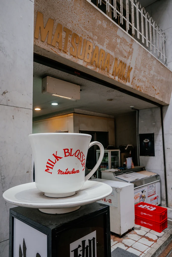 Ingresso del negozio Matsubara Milk lungo Nayamachi Shopping Street con una grande tazza da caffè decorativa bianca in primo piano.