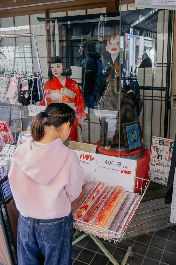 Una bambina di spalle mentre osserva i rotoli di tessuto esposti davanti alla vetrina di un negozio tradizionale con manichini in kimono.