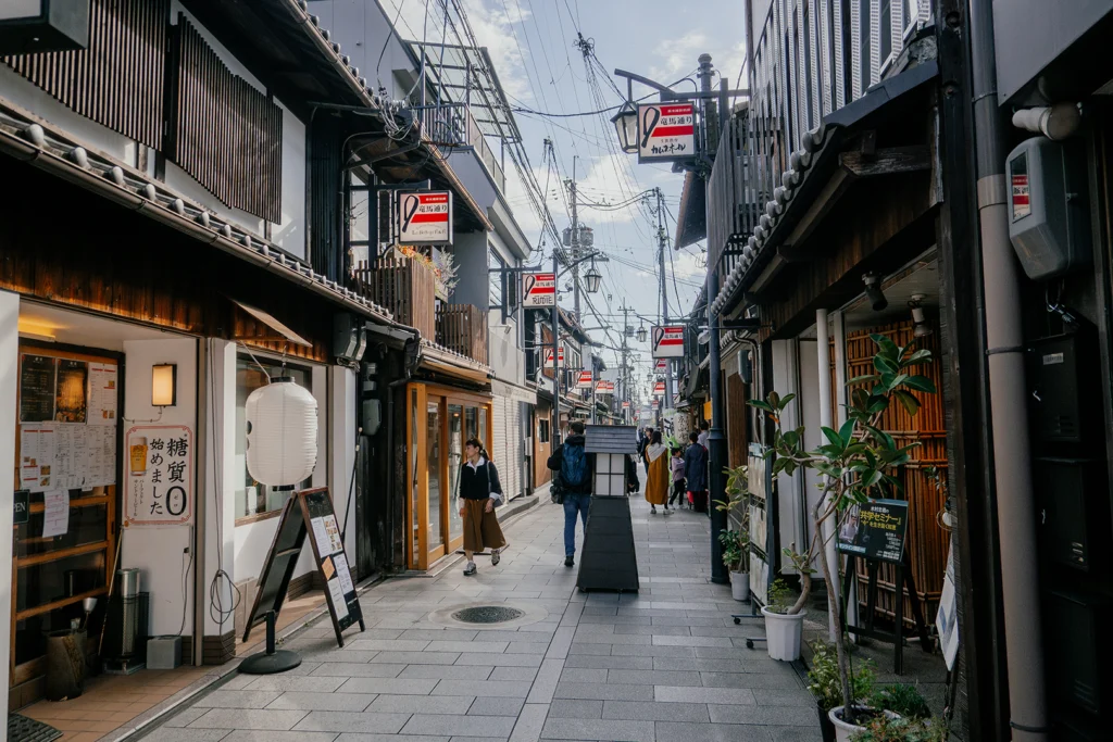 Una veduta prospettica della storica Ryoma-dori Shopping Street a Fushimi, Kyoto, con negozi tradizionali in legno, lanterne bianche appese e persone che passeggiano.