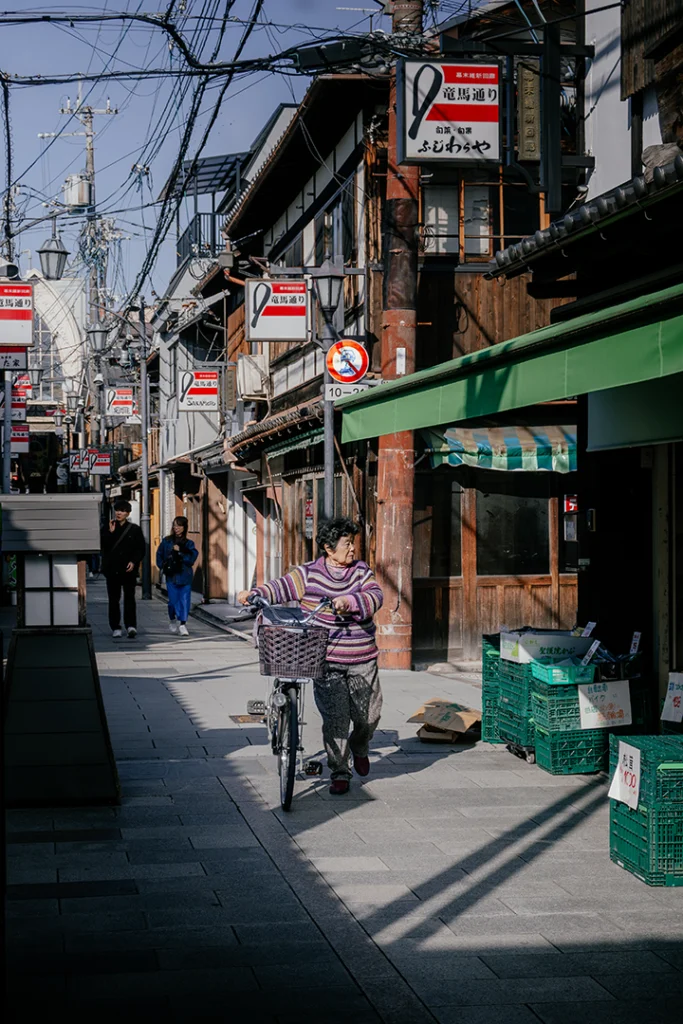 Una signora anziana che spinge una bicicletta lungo la storica Ryoma-dori Shopping Street a Fushimi, Kyoto, tra vecchi edifici in legno ed elementi moderni.