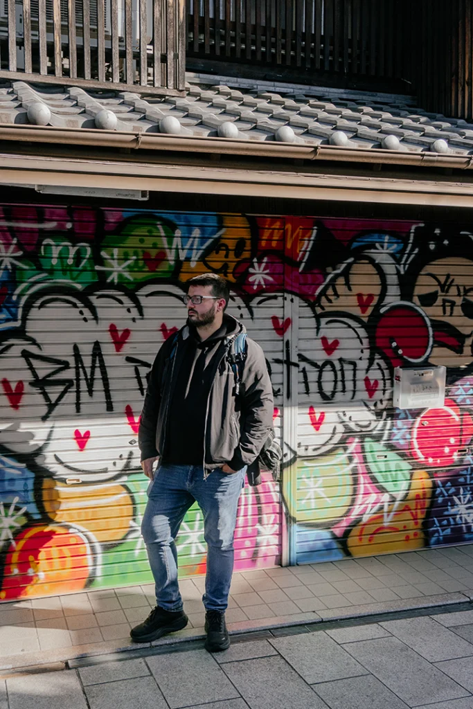 Alfredo di Kappamonogatari posa davanti a una saracinesca coperta di graffiti colorati, sotto il tetto in tegole tradizionali di un edificio a Fushimi, Kyoto.