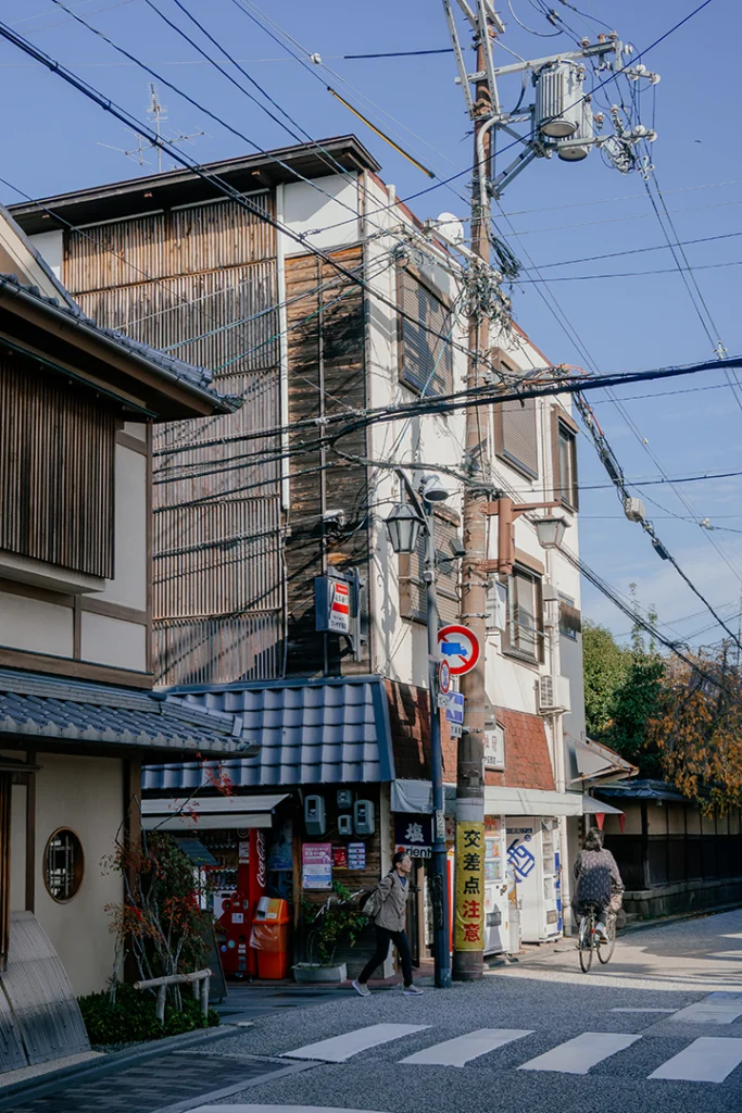 Un incrocio stradale a Fushimi, Kyoto, con strisce pedonali in primo piano, edifici tradizionali e moderni, pali della luce con molti cavi elettrici e passanti.