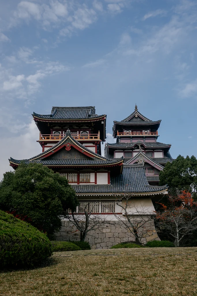 La torre principale e la torre minore del castello di Fushimi Momoyama a Kyoto, viste dal giardino sottostante e incorniciate da foglie di acero autunnali in primo piano.