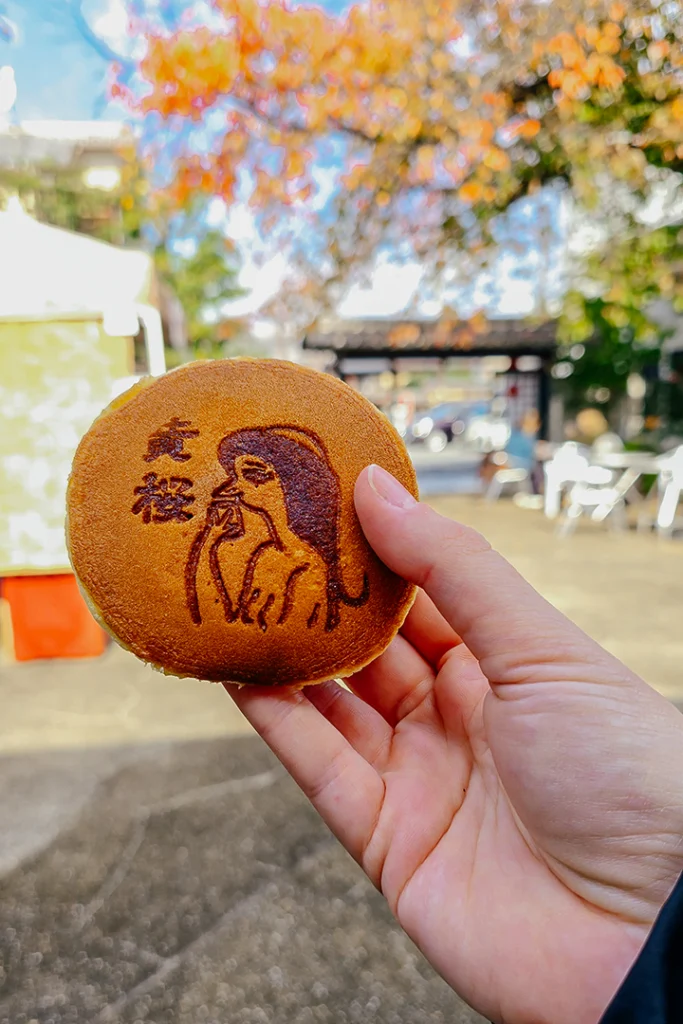 Un dorayaki giapponese rotondo marchiato a fuoco con l'illustrazione di un kappa e scritte in kanji, tenuto in mano all'aperto con alberi autunnali sfocati sullo sfondo.